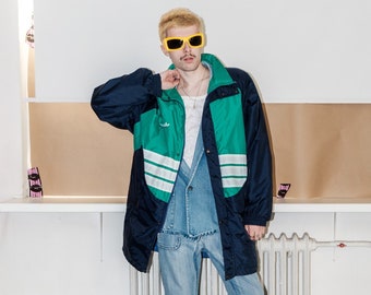 Könnte beinhalten: Eine Person trägt eine blaue und grüne Jacke mit weißen Streifen, hellblaue Jeans und eine gelbe Sonnenbrille. Die Jacke hat einen Reißverschluss und einen Druckknopfverschluss.