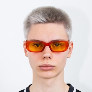 May include: A young person with short, light grey hair wearing a black and white striped shirt and orange rectangular sunglasses.