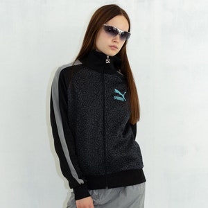 Puede incluir: Una chaqueta negra y gris con cremallera con un logotipo azul de Puma en el pecho izquierdo. La chaqueta tiene un diseño a rayas gris y negro en las mangas. La modelo lleva una falda gris y botas plateadas.