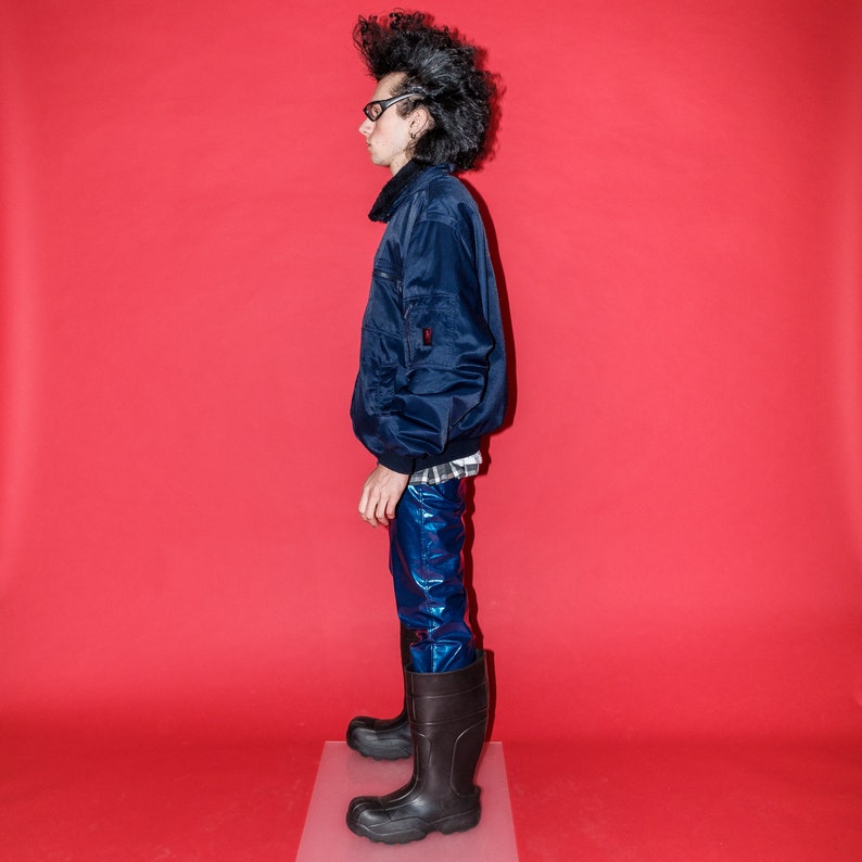 May include: A navy blue bomber jacket with a black collar, paired with shiny blue pants and black rubber boots. The outfit is set against a vibrant red backdrop, creating a bold contrast.