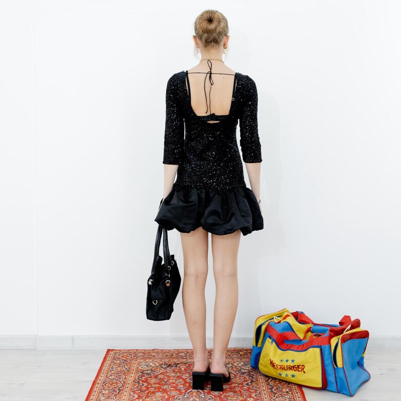 May include: A woman wearing a black sequined dress with a low back and a ruffled hem. She is holding a black handbag and a yellow, red, and blue duffel bag with the text "Meschurger" on it.