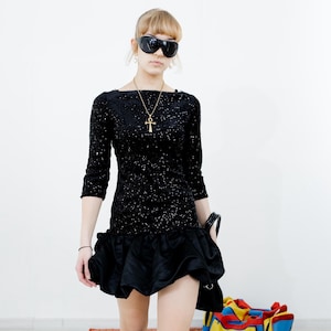 May include: A woman wearing a black sequined dress with a ruffled hem and a gold ankh pendant necklace. She is wearing black sunglasses and holding a black handbag.