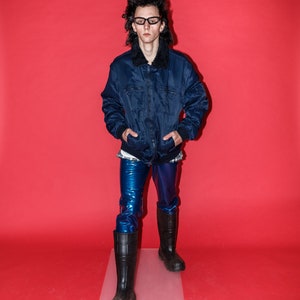 May include: A person wearing a navy blue jacket with a black fur collar, blue shiny pants, black boots, and glasses. The background is a solid red.