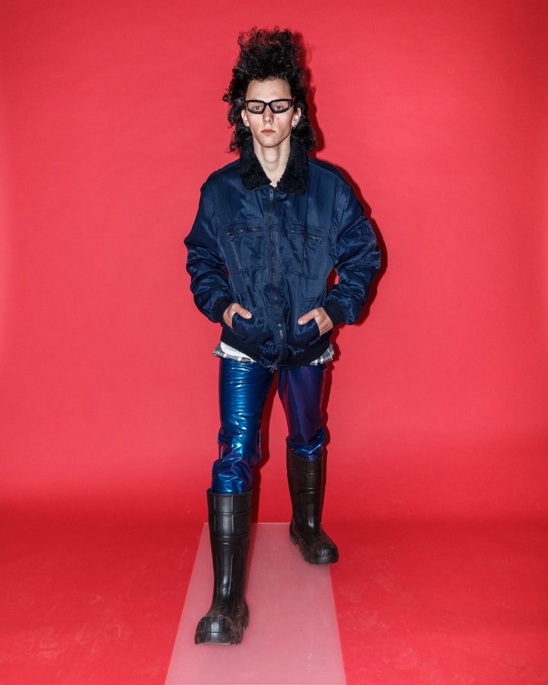 May include: A person wearing a navy blue jacket with a black fur collar, blue shiny pants, black boots, and glasses. The background is a solid red.