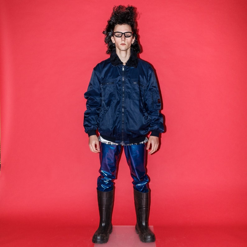 May include: A person wearing a navy blue bomber jacket with a black fur collar, shiny blue pants, and black rubber boots. The background is a solid red.
