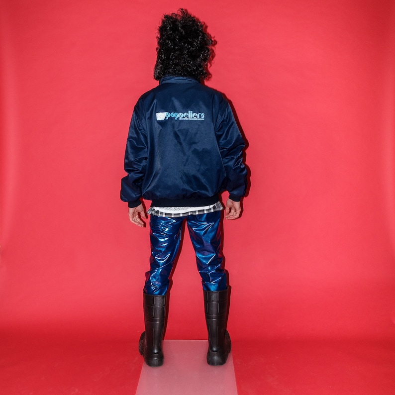 May include: A person wearing a navy blue satin bomber jacket with the word "poppeliers" printed on the back in white and blue. The person is also wearing shiny blue pants, a white mesh shirt, a black and white plaid shirt, and black boots. The background is red.