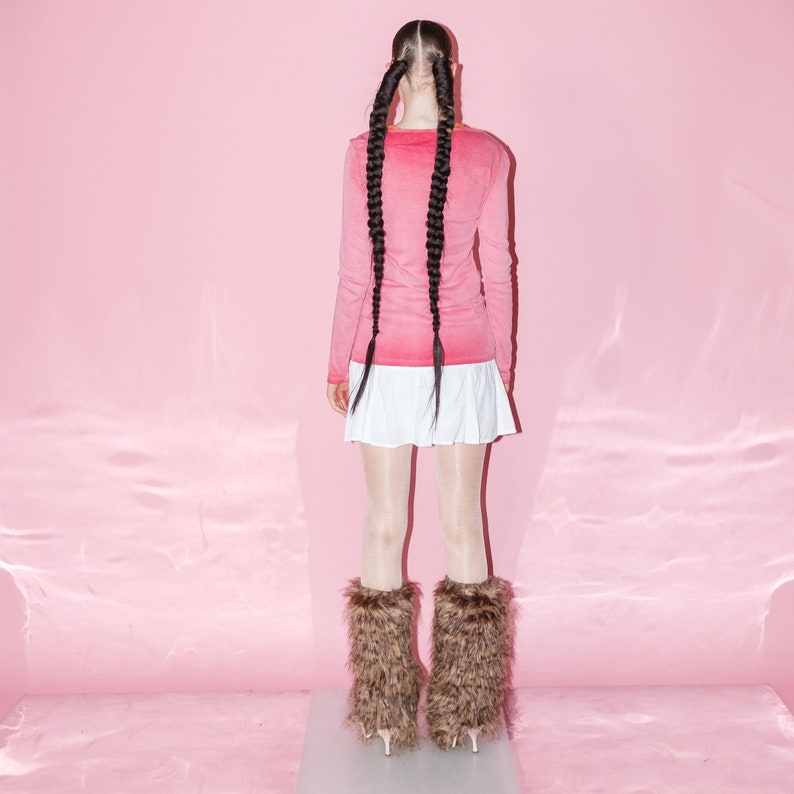 May include: A woman wearing a pink long-sleeved shirt, a white skirt, and brown fur leg warmers. She is standing in front of a pink background.
