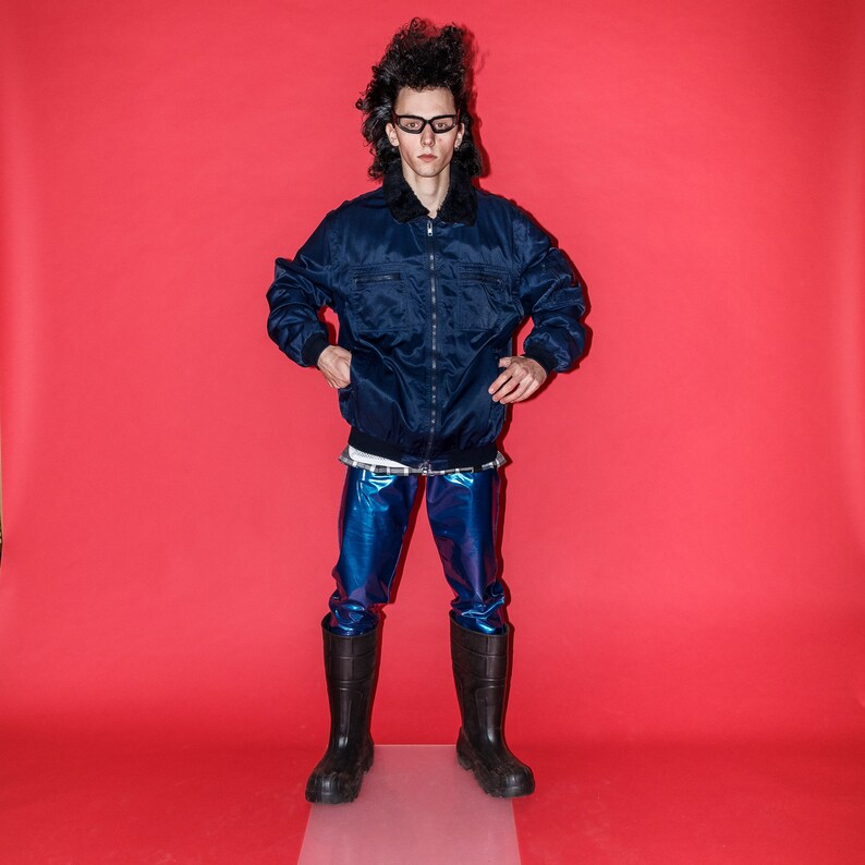 May include: A person wearing a navy blue bomber jacket with a black fur collar, blue metallic pants, and black rubber boots. The background is a solid red.