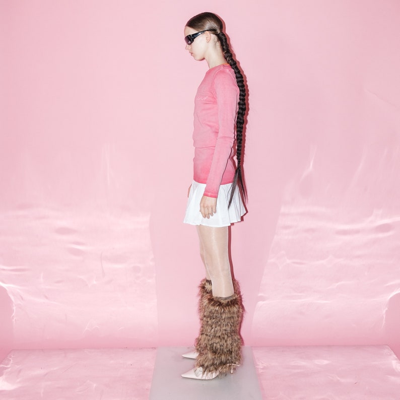 May include: A woman wearing a pink long-sleeved sweater, a white pleated skirt, and brown fur leg warmers. She is standing on a pink background and wearing sunglasses.