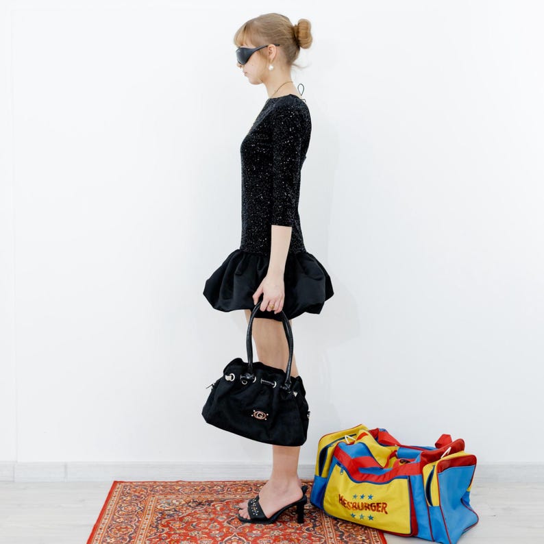 May include: A woman wearing a black sparkly dress with a ruffled hem, black sunglasses, and black heeled shoes. She is holding a black handbag and standing in front of a white wall with a red, yellow, and blue travel bag with the text "MESSBURGER" on it.