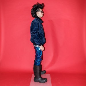 May include: A person wearing a navy blue bomber jacket, shiny blue pants, and black rubber boots. The jacket has a black collar and a small pocket on the chest. The boots are knee-high and have a thick sole. The background is a solid red.
