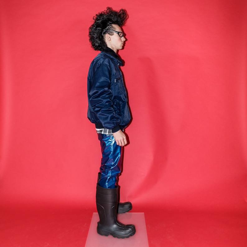 May include: A person wearing a navy blue bomber jacket, shiny blue pants, and black rubber boots. The jacket has a black collar and a small pocket on the chest. The boots are knee-high and have a thick sole. The background is a solid red.