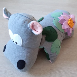 Könnte beinhalten: Ein grauer Plüsch-Nilpferd mit einem rosa Ohr und einer schwarzen Nase. Das Nilpferd liegt auf der Seite mit dem Kopf auf dem Boden. Das Nilpferd hat eine grüne und rosa Blume auf dem Rücken.