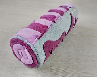 Puede incluir: Un juguete cilíndrico para mascotas en tonos grises, morados y lavanda. El juguete presenta un diseño en espiral en un extremo y está adornado con varias formas y rayas de tela. El material suave y afelpado está diseñado para el juego interactivo.