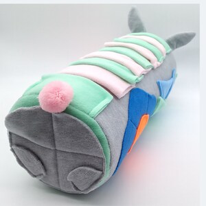 Op de afbeelding: Een grijs pluche speelgoed in de vorm van een konijn met een roze pomponstaart. Het konijn is gemaakt van verschillende gekleurde stoffen, waaronder groen, blauw en oranje.