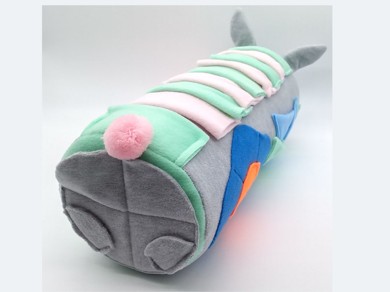Op de afbeelding: Een grijs pluche speelgoed in de vorm van een konijn met een roze pomponstaart. Het konijn is gemaakt van verschillende gekleurde stoffen, waaronder groen, blauw en oranje.