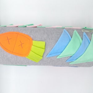 Op de afbeelding: Een grijs en wit cilindrisch kussen met een viltwortel, groene bladeren en blauwe en groene vilt driehoeken erop genaaid. Het kussen is gemaakt van zachte stof en is perfect voor een kinderkamer.