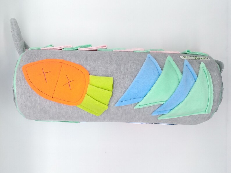 Op de afbeelding: Een grijs en wit cilindrisch kussen met een viltwortel, groene bladeren en blauwe en groene vilt driehoeken erop genaaid. Het kussen is gemaakt van zachte stof en is perfect voor een kinderkamer.