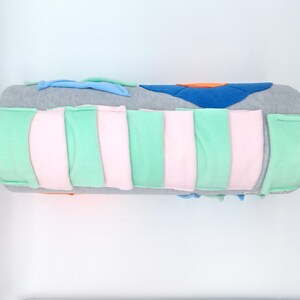 Op de afbeelding: Een grijs en wit zintuiglijk speelgoed voor baby's en peuters. Het speelgoed heeft de vorm van een cilinder en heeft verschillende texturen en kleuren. Het heeft een roze pompon aan het ene uiteinde.