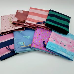Könnte beinhalten: Eine Sammlung von acht Stoffbeuteln in verschiedenen Farben und Mustern. Designs umfassen rosa Sterne, rosa Punkte, rosa und weiße Streifen, grün und marineblaue Streifen, blaue Wolken, lila Regentropfen und ein rosa-blaues Batikmuster. Jeder Beutel hat einen Kordelzugverschluss.