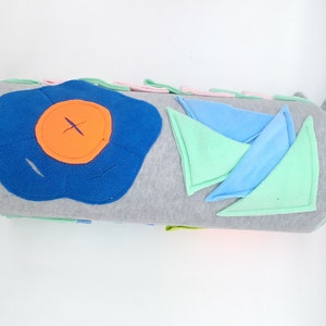 Op de afbeelding: Een grijs en wit cilindrisch kussen met verschillende gekleurde stoflapjes die erop zijn genaaid. De lapjes omvatten een blauwe bloem met een oranje middelpunt, groene en blauwe driehoeken en een roze rechthoek.