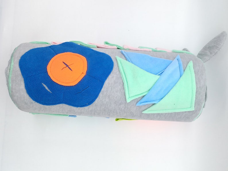 Op de afbeelding: Een grijs en wit cilindrisch kussen met verschillende gekleurde stoflapjes die erop zijn genaaid. De lapjes omvatten een blauwe bloem met een oranje middelpunt, groene en blauwe driehoeken en een roze rechthoek.
