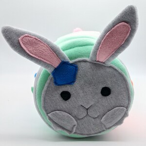 Op de afbeelding: Een pluche speelgoed in de vorm van een konijn met een grijs gezicht, roze oren en een groen-wit gestreept lichaam. Het konijn heeft een blauwe ster-patch op zijn hoofd en zwarte ogen.
