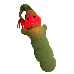 Puede incluir: Un peluche verde con forma de oruga con una cara sonriente de color rojo brillante y ojos azules. Tiene un sombrero largo y verde y zarcillos naranjas. El cuerpo está segmentado, creando un juguete suave y caprichoso.