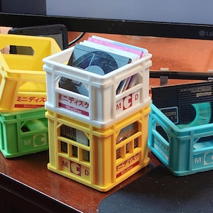Op de afbeelding: Een stapel kleurrijke, kleine plastic kratten, elk met de letters "MCD" en Japanse tekst. De kratten zijn geel, groen, wit en blauw en bevatten mini-discs. De kratten staan op een bruin oppervlak.