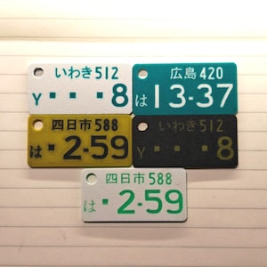May include: A collection of rectangular Japanese license plate tags in various colors: white, teal, yellow, and black. Each tag features Japanese characters and numbers, with the text "いわき 512", "広島 420", and "四日市 588" visible.