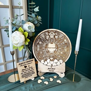 May include: A wooden guest book with a circular design featuring an Iron Man character and other Marvel characters. The guest book is on a stand with a sign that says "Guestbook Please Sign A & Drop It Into The Frame So We Will Always Remember The Loved Ones Who Came" and "Adam & Bechn 19-08-2023".