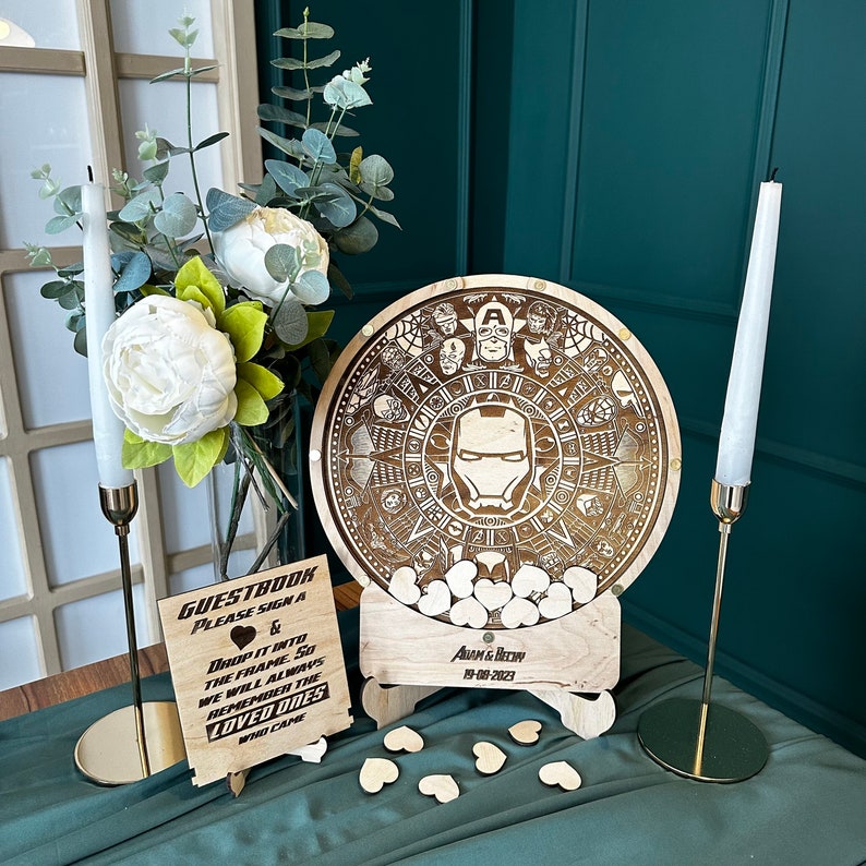 May include: A wooden guest book with a circular design featuring an Iron Man character and other Marvel characters. The guest book is on a stand with a sign that says "Guestbook Please Sign A & Drop It Into The Frame So We Will Always Remember The Loved Ones Who Came" and "Adam & Bechn 19-08-2023".