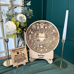 May include: A wooden guest book with a circular design featuring a superhero theme. The design includes a central image of Iron Man's mask and other superhero symbols. The guest book is on a wooden stand with a sign that says "GUESTBOOK PLEASE SIGN & DROP IT INTO THE FRAME SO WE WILL ALWAYS REMEMBER THE LOVED ONES WHO CAME" and "ADAM & BEY 19-08-2023".