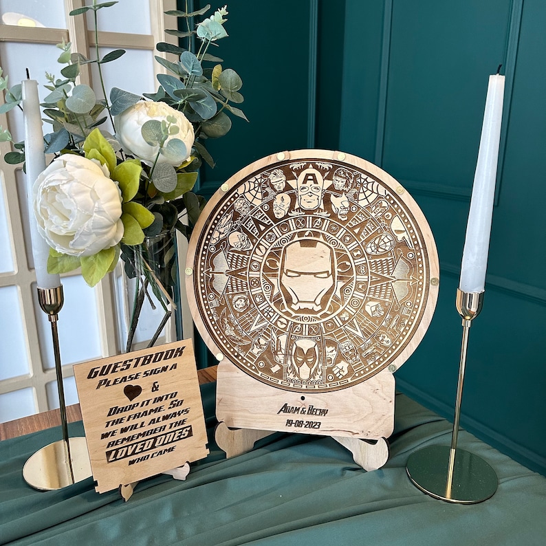 May include: A wooden guest book with a circular design featuring a superhero theme. The design includes a central image of Iron Man's mask and other superhero symbols. The guest book is on a wooden stand with a sign that says "GUESTBOOK PLEASE SIGN & DROP IT INTO THE FRAME SO WE WILL ALWAYS REMEMBER THE LOVED ONES WHO CAME" and "ADAM & BEY 19-08-2023".