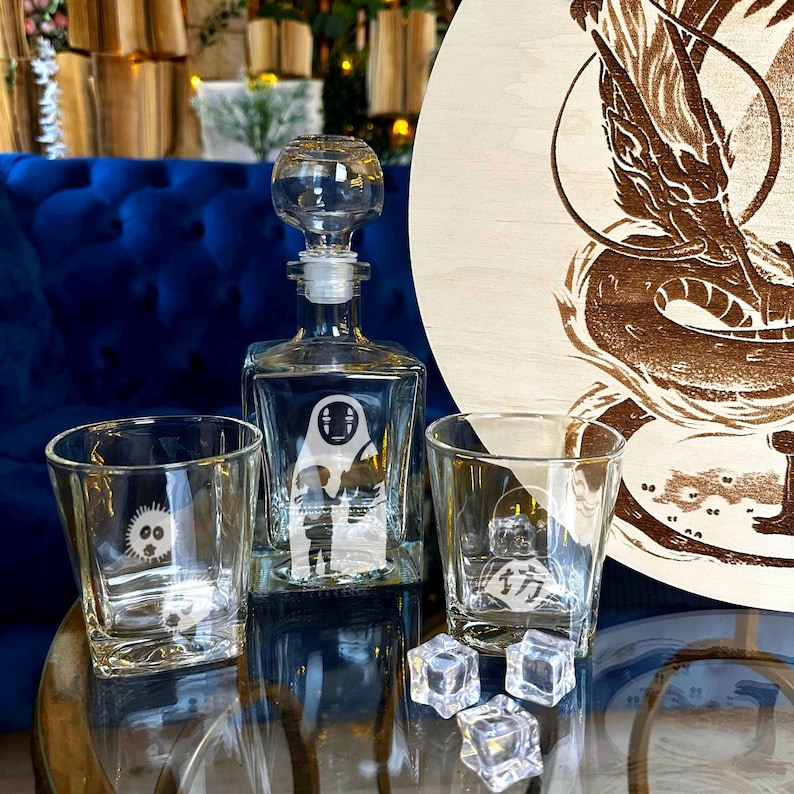 May include: A clear glass decanter with a stopper, two square glasses, and a round wooden board with a dragon design. The decanter and glasses feature etched character designs. Clear ice cubes are in front of the glasses.