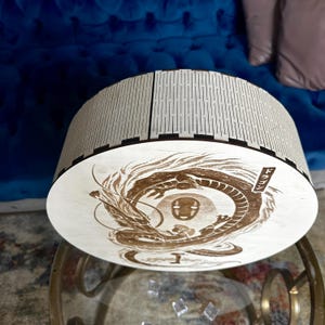 May include: A round, wooden box with a laser-etched design of a dragon and a character from a Japanese anime. The box has a light wood finish with a decorative, slotted top. The dragon design is in shades of brown.