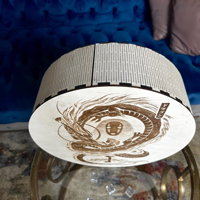May include: A round, wooden box with a laser-etched design of a dragon and a character from a Japanese anime. The box has a light wood finish with a decorative, slotted top. The dragon design is in shades of brown.