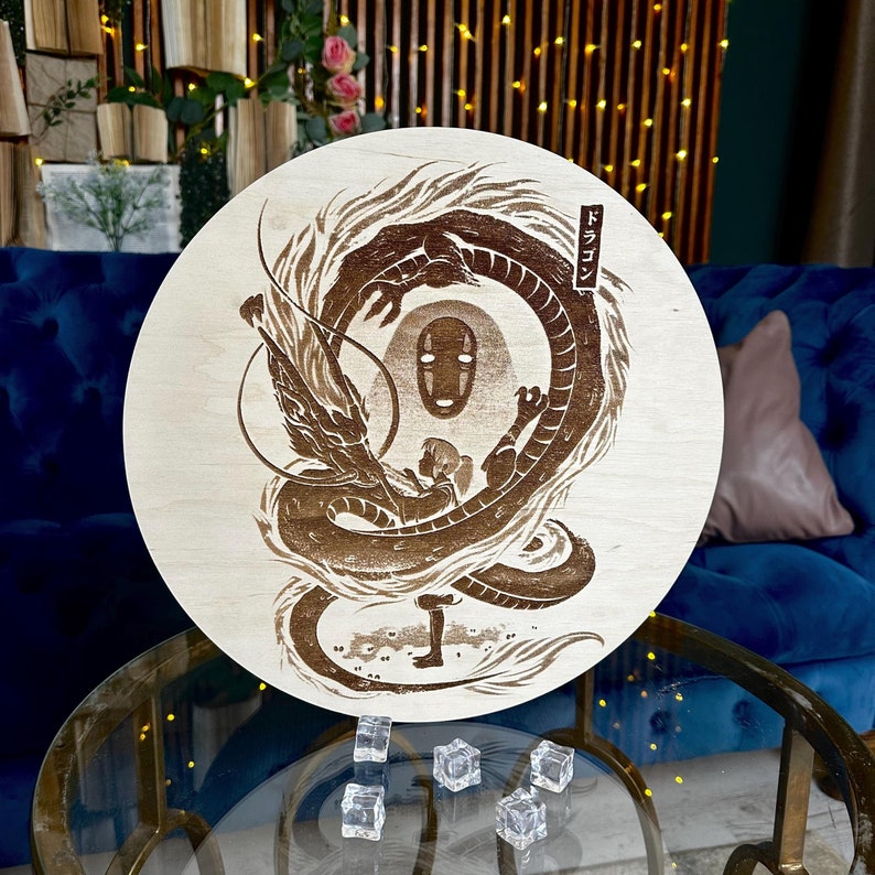 May include: A round, light-colored wooden plaque featuring a detailed brown illustration of a dragon, a figure, and a character with a mask. Japanese text is visible in the upper right corner. The plaque is displayed on a glass table.