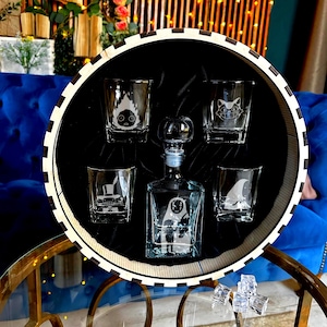 May include: A clear glass decanter and shot glasses set inside a round tray. The decanter and shot glasses have etched character designs. The tray has a black interior and a white and black striped rim. The set is displayed on a glass table.