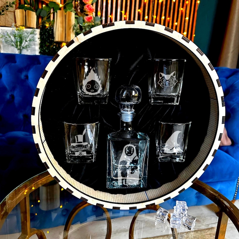 May include: A clear glass decanter and shot glasses set inside a round tray. The decanter and shot glasses have etched character designs. The tray has a black interior and a white and black striped rim. The set is displayed on a glass table.