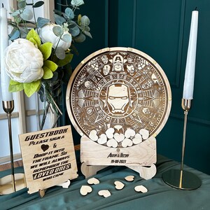 May include: A wooden guest book box with a circular wooden top featuring a laser-engraved design of Iron Man and other Marvel characters. The box is filled with small wooden hearts and has a sign that reads "GUESTBOOK PLEASE SIGN A & DROP IT INTO THE FRAME SO WE WILL ALWAYS REMEMBER THE LOVED ONES WHO CAME" and "ADAM & BECHY 19-08-2023".