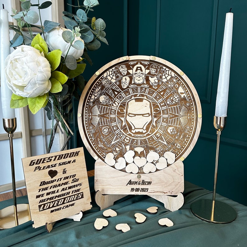May include: A wooden guest book box with a circular wooden top featuring a laser-engraved design of Iron Man and other Marvel characters. The box is filled with small wooden hearts and has a sign that reads "GUESTBOOK PLEASE SIGN A & DROP IT INTO THE FRAME SO WE WILL ALWAYS REMEMBER THE LOVED ONES WHO CAME" and "ADAM & BECHY 19-08-2023".