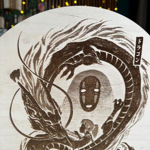 May include: A wooden round featuring a detailed, brown laser-etched design from a Japanese animated film. The artwork depicts a dragon, a faceless spirit, and a young girl. Japanese text is visible on the right side of the design.