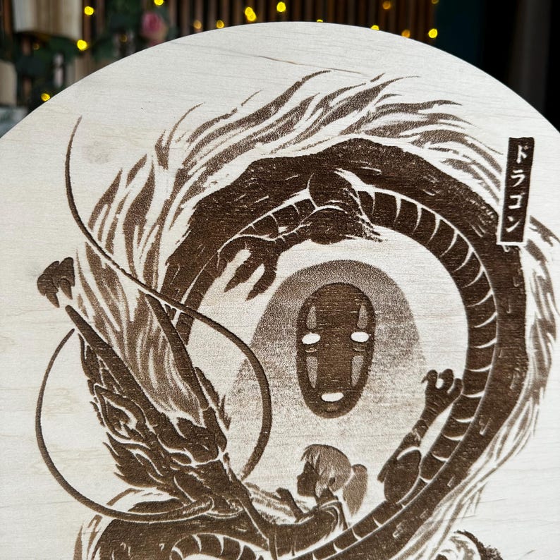 May include: A wooden round featuring a detailed, brown laser-etched design from a Japanese animated film. The artwork depicts a dragon, a faceless spirit, and a young girl. Japanese text is visible on the right side of the design.