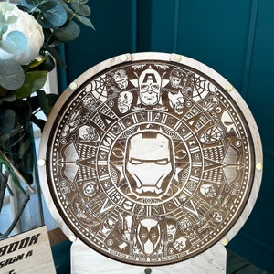 May include: A wooden round wall clock with a detailed design featuring a compass, stars, and various superhero characters. The clock face is surrounded by a ring of superhero characters, including Captain America, Iron Man, and Thor. The clock is made of wood and has a brown finish.