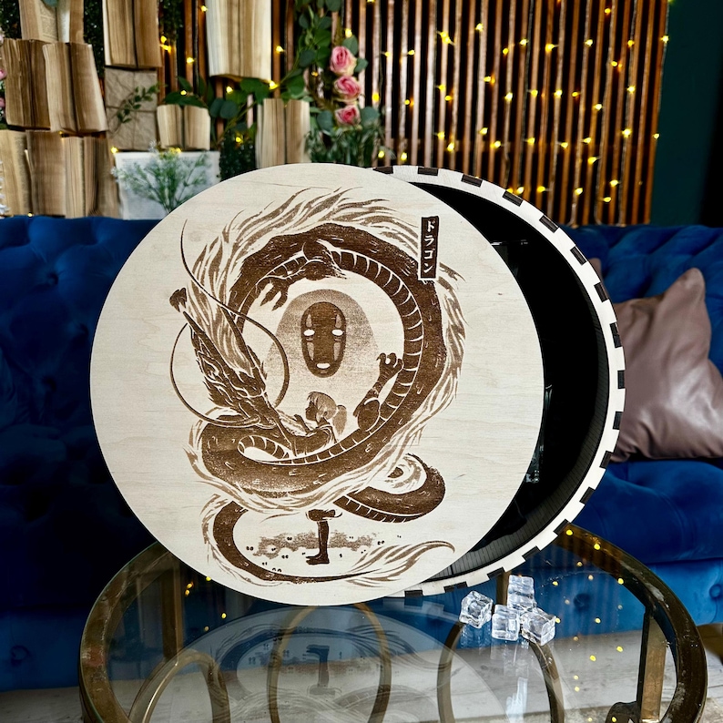 May include: A round wooden box with a detailed brown engraving of a dragon, a figure, and a character from a Japanese animated film. The box has a black interior and a white and black striped edge. Japanese text is visible.