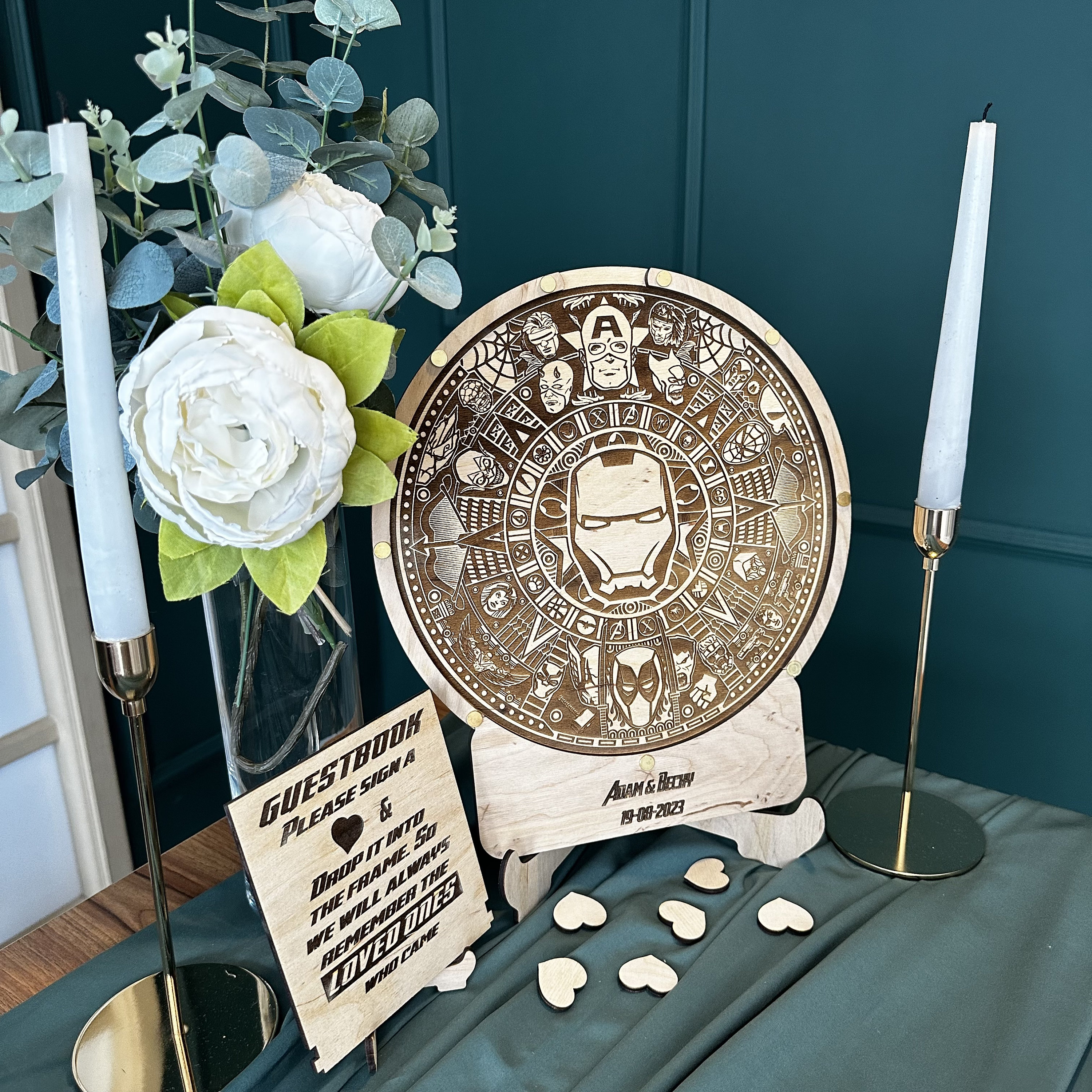 May include: A wooden guest book sign with a heart and the text "GUESTBOOK PLEASE SIGN & DROP IT INTO THE FRAME SO WE WILL ALWAYS REMEMBER THE LOVED ONES WHO CAME". A wooden round sign with a design of various Marvel characters, including Iron Man, Captain America, and Spider-Man. The sign says "Adam & Becky 19-08-2023".