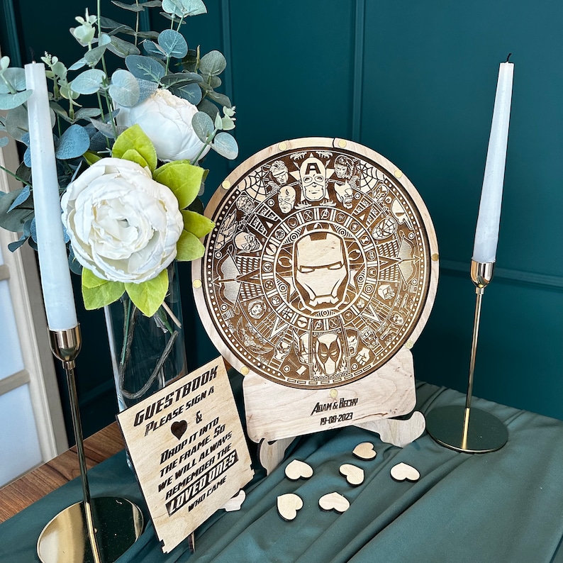 May include: A wooden guest book sign with a heart and the text "GUESTBOOK PLEASE SIGN & DROP IT INTO THE FRAME SO WE WILL ALWAYS REMEMBER THE LOVED ONES WHO CAME". A wooden round sign with a design of various Marvel characters, including Iron Man, Captain America, and Spider-Man. The sign says "Adam & Becky 19-08-2023".