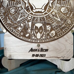May include: A wooden circular plaque with a laser-engraved design featuring various Marvel characters, including Deadpool, Iron Man, and Captain America. The plaque is mounted on a wooden stand with the text "ADAM & BECKY 19-08-2023" engraved on the front.