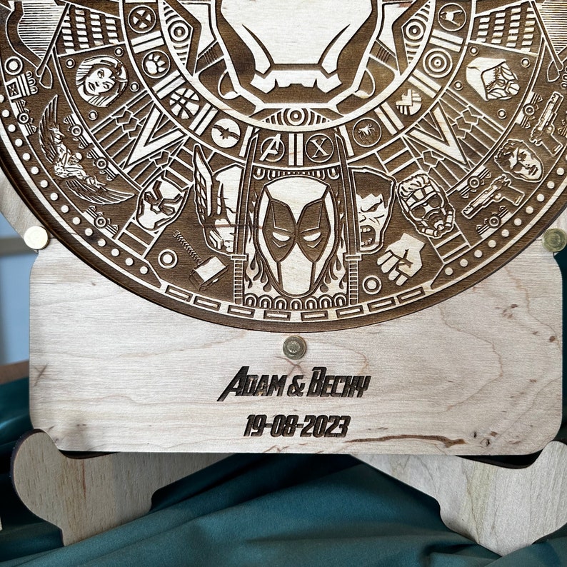 May include: A wooden circular plaque with a laser-engraved design featuring various Marvel characters, including Deadpool, Iron Man, and Captain America. The plaque is mounted on a wooden stand with the text "ADAM & BECKY 19-08-2023" engraved on the front.